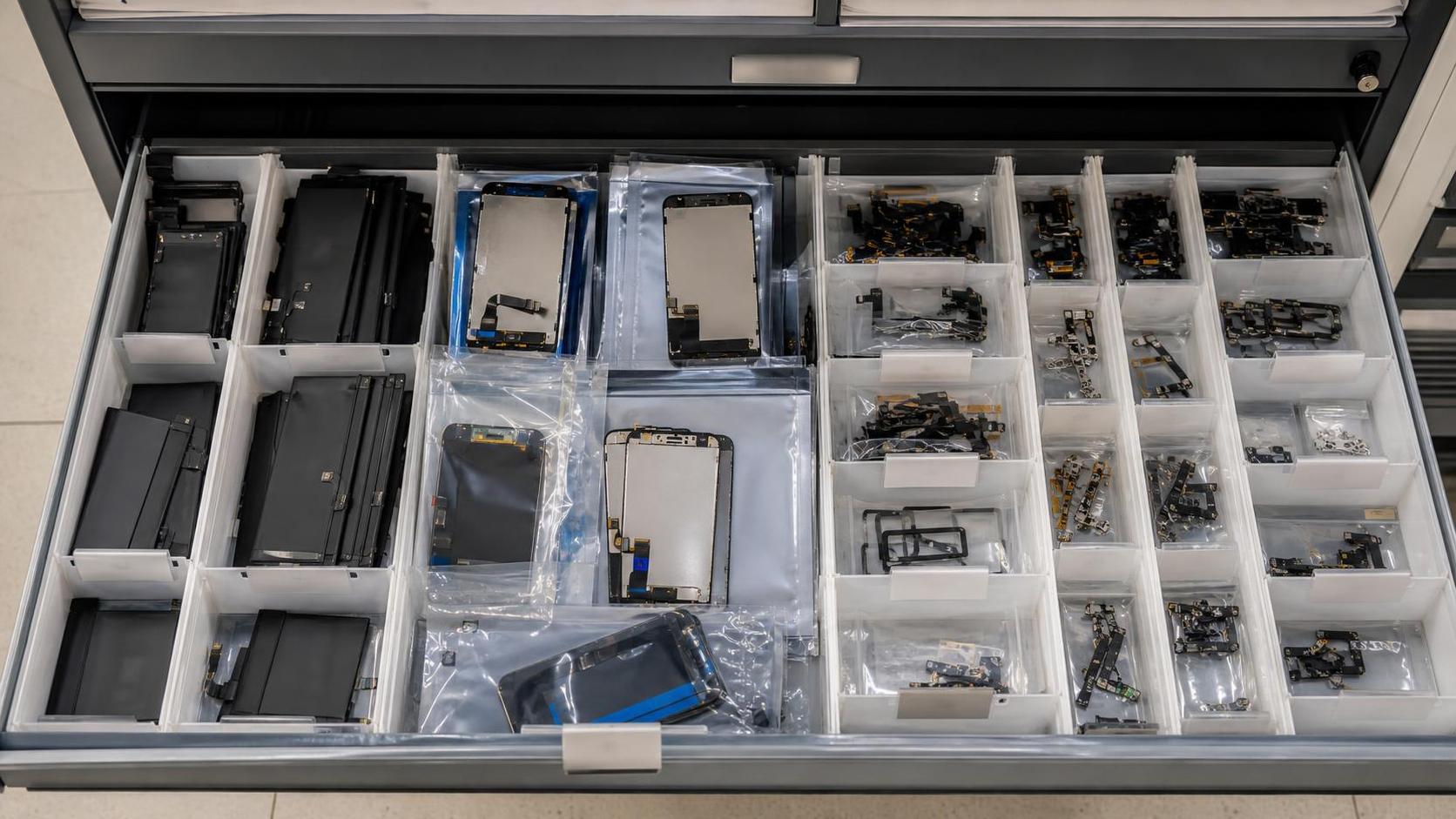 Cajón de taller con baterías, pantallas y piezas de smartphone organizadas para reparación